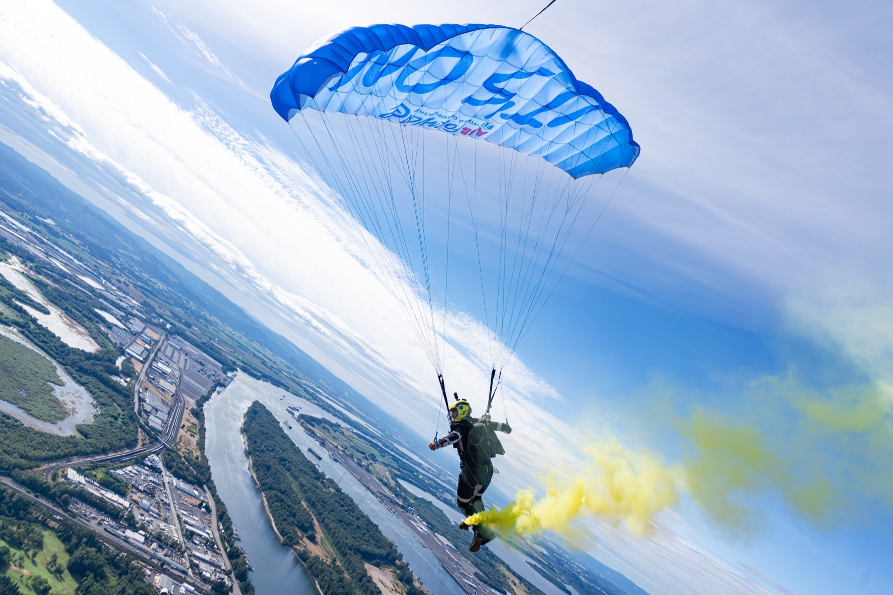 Skydiver under a blue canopy trailing yellow smoke above a river
